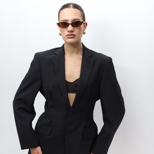 May include: A black pinstripe blazer with a tailored fit. The blazer features a single-button closure and front pockets. The model is wearing brown sunglasses and gold earrings. The outfit is completed with a black bra.