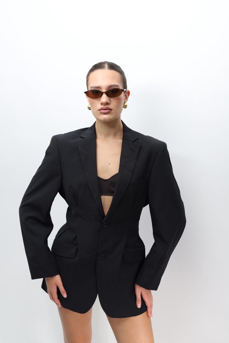 May include: A black pinstripe blazer with a tailored fit. The blazer features a single-button closure and front pockets. The model is wearing brown sunglasses and gold earrings. The outfit is completed with a black bra.