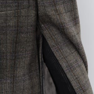 May include: Close-up of a tweed blazer in brown, grey, and blue hues. The fabric features a plaid design. The interior lining is black. A pocket of the blazer is visible.