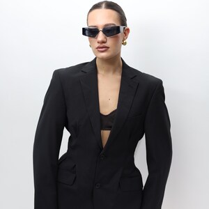 May include: A black pinstripe blazer with a two-button closure and notched lapels. The blazer is paired with black sunglasses and gold earrings. The outfit is completed with a black bra. The background is plain white.