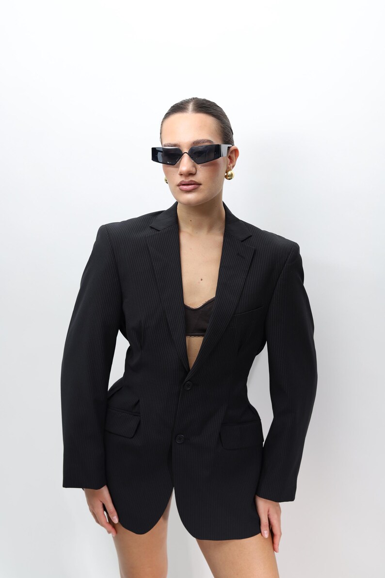 May include: A black pinstripe blazer with a two-button closure and notched lapels. The blazer is paired with black sunglasses and gold earrings. The outfit is completed with a black bra. The background is plain white.