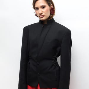 Puede incluir: Un blazer negro con cuello mandarín y mangas largas. El blazer presenta un detalle único de cordones en los laterales y se combina con pantalones rojos. El conjunto se presenta sobre un fondo blanco.