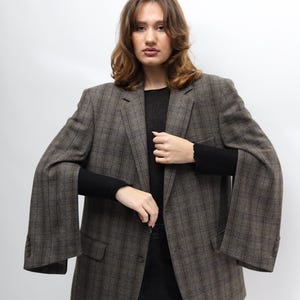 May include: A brown plaid blazer, black top and black trousers. The blazer features a classic design with a notched lapel and button closure. The sleeves are long and wide. The outfit is set against a white background.