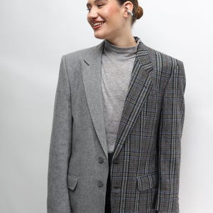 Pode incluir: Um blazer de duas cores, com um lado cinza liso e o outro com um padrão xadrez cinza e azul. O blazer tem uma lapela entalhada clássica e fecho de botão. A modelo está a usar uma gola alta cinzenta e uma saia preta.