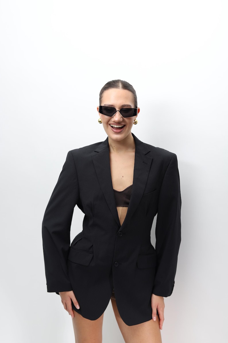 May include: A black pinstripe blazer with a tailored fit. The blazer features a notched lapel, two front pockets, and a single-button closure. The model is wearing black sunglasses and gold earrings. The blazer is a classic piece of apparel.