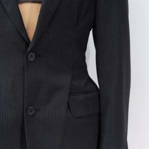 May include: Black pinstripe blazer with two-button closure and a notched lapel. The blazer features a single chest pocket and two front pockets. The fabric has a subtle vertical stripe pattern. The sleeves are long and the fit appears tailored.