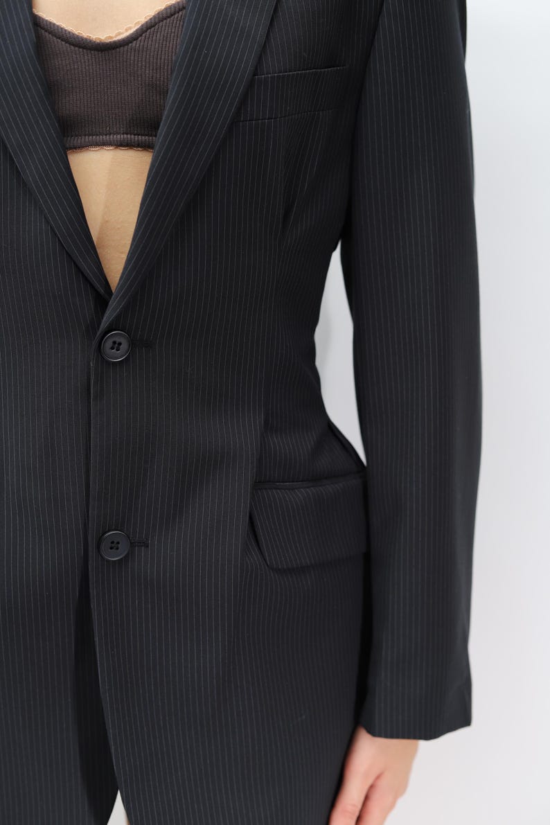 May include: Black pinstripe blazer with two-button closure and a notched lapel. The blazer features a single chest pocket and two front pockets. The fabric has a subtle vertical stripe pattern. The sleeves are long and the fit appears tailored.