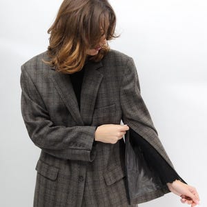 May include: A brown plaid blazer with a classic design. The blazer features a notched lapel, front pockets, and a button closure. The sleeves are long and the interior lining is black. The blazer is worn over a black top.