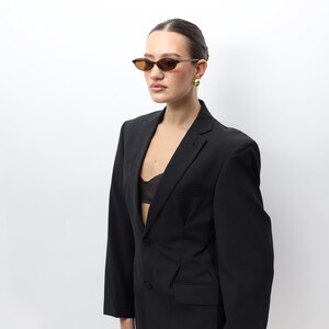 May include: A black pinstripe blazer with a two-button closure. The blazer is paired with brown-tinted sunglasses and gold earrings. The outfit is set against a white background.