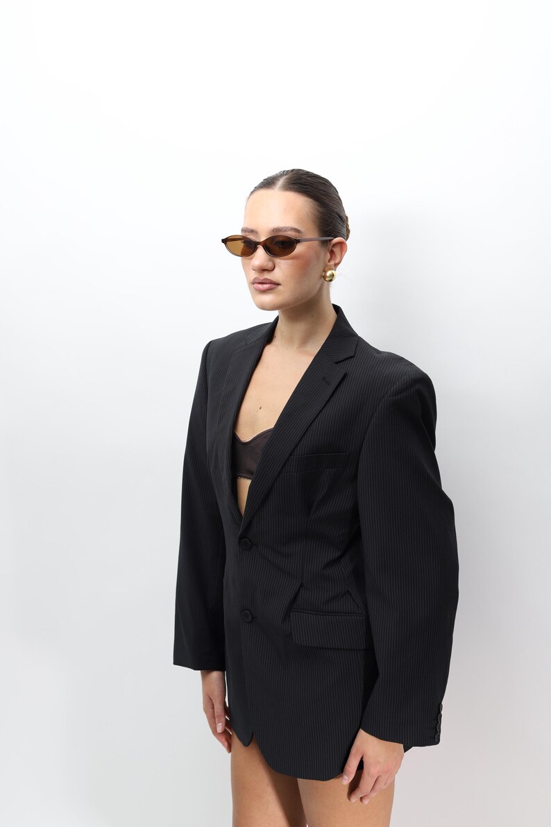 May include: A black pinstripe blazer with a two-button closure. The blazer is paired with brown-tinted sunglasses and gold earrings. The outfit is set against a white background.