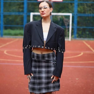 May include: A person wearing a black cropped blazer with silver rings on the sleeves and a black and white plaid skirt. The blazer has a white undershirt visible.