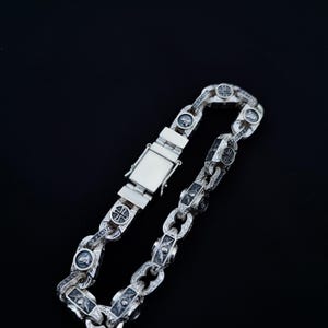 Puede incluir: Un brazalete de cadena de plata con un diseño detallado que presenta pequeñas cruces y figuras. El brazalete tiene un cierre de plata.