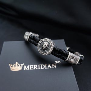 Puede incluir: Un brazalete de cuero negro con un dije de plata que presenta una cara y el texto "925 MERIDIAN".
