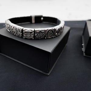 Puede incluir: Un brazalete de plata con una banda de cuero negro y múltiples símbolos nórdicos grabados, incluyendo el Valknut, Mjolnir y Vegvisir. El brazalete se muestra en una caja negra.