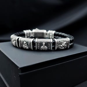 Puede incluir: Una pulsera de plata con una banda de cuero trenzado negro. La pulsera presenta una iconografía religiosa detallada en eslabones rectangulares de plata. El cierre también es plateado. La pulsera se exhibe en una caja negra sobre un fondo oscuro.