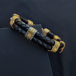 Puede incluir: Pulsera de cuero trenzado negro con detalles dorados. La pulsera presenta bandas decorativas doradas con intrincados diseños. El diseño incluye una trenza de doble hebra, creando un accesorio elegante.