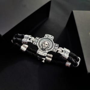 Puede incluir: Un brazalete de cuero negro con un colgante de cruz de plata que presenta una imagen detallada de Jesús. La cruz tiene letras cirílicas alrededor del borde.