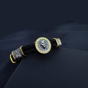 Puede incluir: Un brazalete de cuero negro con un colgante de plata y oro que presenta una imagen detallada de Jesucristo. El colgante está rodeado por un borde dorado con un patrón decorativo.