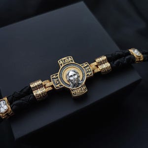 Puede incluir: Un brazalete de cuero negro con un colgante de cruz dorada que presenta una imagen plateada de Jesús. La cruz tiene letras cirílicas.