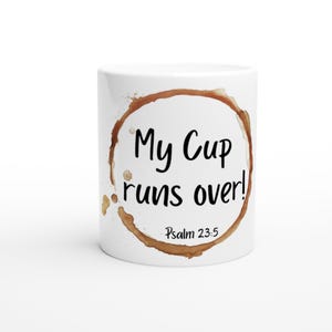 Puede incluir: Taza de café de cerámica blanca con una mancha de café marrón. La taza tiene texto negro que dice "My Cup runs over! Psalm 23:5".