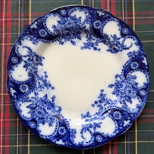 May include: An antique white and blue plate featuring a floral design. The plate has a scalloped edge and a detailed blue floral pattern around the rim. The centre of the plate is white.