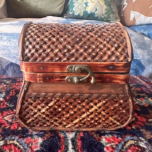 May include: A small, decorative chest with a woven, basket-like design. The chest is made of brown wood and has a hinged lid with a metal clasp. The chest sits on a patterned rug, suggesting a vintage or antique aesthetic.