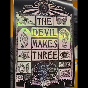May include: Black and holographic print featuring the text "THE DEVIL MAKES THREE" with various illustrations. Includes a skull, hand, butterfly, mushroom, eye, flower, and insect. Also includes the cities of Denver, Colorado Springs, Steamboat Springs, Fort Collins, and Boulder.