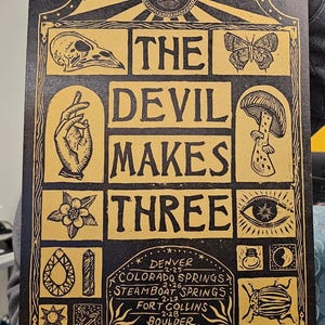 May include: A black and gold print with the text "THE DEVIL MAKES THREE" and various illustrations. The print includes a skull, butterfly, hand, mushroom, flower, eye, gem, and beetle. It also lists Denver, Colorado Springs, Steamboat Springs, Fort Collins, and Boulder.