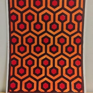 May include: A print of the iconic Overlook Hotel carpet pattern from the movie "The Shining". The pattern features a repeating design of red and orange hexagons on a black background. The print is signed and numbered.