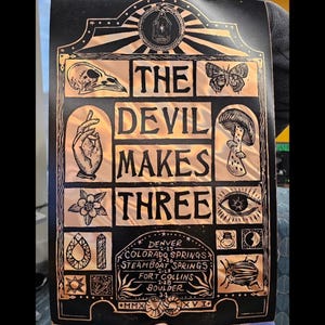 May include: A black and copper-colored art print with the text "THE DEVIL MAKES THREE". The print features various illustrations, including a skull, butterfly, mushroom, eye, and a hand. It also includes a list of tour dates for Denver, Colorado Springs, Steamboat Springs, Fort Collins, and Boulder.