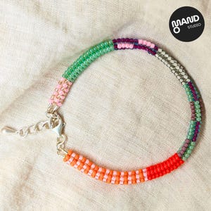 Può includere: Un braccialetto colorato con perline e chiusura argentata. Il braccialetto presenta sezioni di perline verdi, rosa, arancioni e rosse. Il braccialetto è su uno sfondo di tessuto color neutro. Il logo del marchio è in alto a destra.