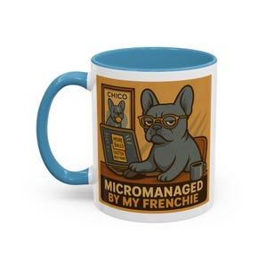 Pode incluir: Caneca de cerâmica branca com borda azul. A caneca apresenta uma ilustração de desenho animado de um bulldogue francês usando óculos e sentado em uma mesa com um laptop. O texto "Micromanaged by my Frenchie" está impresso na caneca.