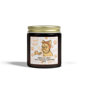 Puede incluir: Un frasco de vidrio marrón con tapa dorada, con una ilustración humorística de un bulldog con pañuelo rojo y el texto "Smells Like: You Touched Another Dog". La etiqueta incluye huellas de patas y huesos.