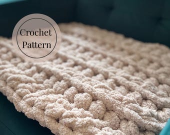 Può includere: Primo piano di una coperta all'uncinetto beige con un design grosso e strutturato. La coperta è piegata su una superficie turchese. Le parole "Crochet Pattern" sono in un grafico circolare.