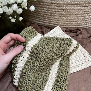 May include: Handmade crochet washcloths in olive green and cream. One washcloth is folded, displaying a striped pattern, while another lies flat. The textured stitch creates a rustic, cosy feel. Ideal for kitchen or bathroom use.