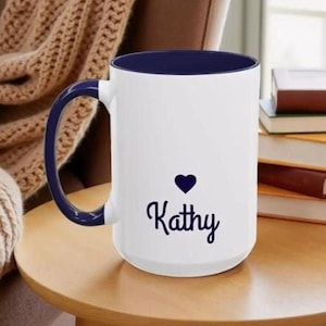 A white mug with a blue rim, featuring the name "Kathy" printed on it, sits on a wooden table.