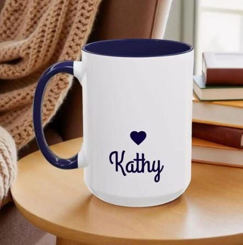 A white mug with a blue rim, featuring the name "Kathy" printed on it, sits on a wooden table.