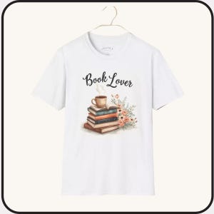 May include: White t-shirt with a watercolor design featuring a stack of books, a coffee cup, and flowers. The words "Book Lover" are written above the design in a script font. The shirt is hanging on a hanger.