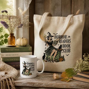 May include: A beige canvas tote bag and a white ceramic mug, both featuring a graphic illustration of a witch reading a book with the text Horror Readers Book Club, accompanied by a black cat, a pumpkin, and small stars on a light background.