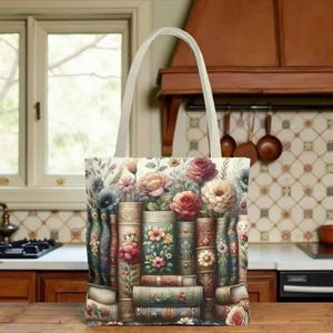 May include: A floral tote bag with a cream-colored handle. The bag features a design of vintage books adorned with flowers in shades of pink, peach, and blue. The background includes additional floral elements, creating a whimsical and artistic aesthetic.