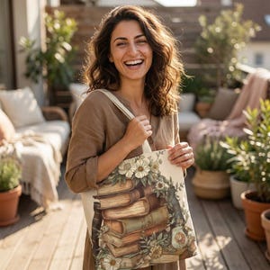 May include: A beige tote bag featuring a stack of vintage books and floral accents. The design includes cream-colored flowers and green foliage. The bag has a light-colored strap and is held by a person on a wooden deck.