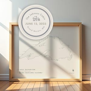 May include: Framed map print of Lake Superior with a light beige background and a wooden frame. The map includes the text "LAKE SUPERIOR" and a circular graphic with the text "The Johnson Cabin JUNE 12, 2025 Grand Marais, MN".