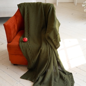 May include: A large, olive green knitted throw blanket featuring a textured cable knit border and a smooth central panel. The blanket is draped over an orange armchair and has a small, round red apple resting on its surface near the edge.