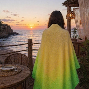Puede incluir: Una persona con un poncho de punto degradado amarillo y verde, de pie en una playa al atardecer. El poncho hace la transición de amarillo a verde. El fondo incluye una mesa de madera, el océano y una pequeña estructura con luces colgantes.