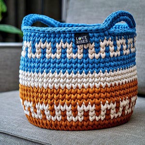 May include: A handmade, round crochet basket with two handles. The basket features a color-blocked design with blue, white, and orange sections. The text "LOVE RISES" is on a small black tag attached to the basket.