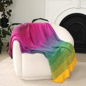 Op de afbeelding: Een kleurrijke deken in roze, paarse, groene en gele tinten, gedrapeerd over een witte fauteuil. De deken heeft een gefranjerde rand. De stoel staat tegen een neutrale achtergrond, wat een gezellige en uitnodigende sfeer creëert.