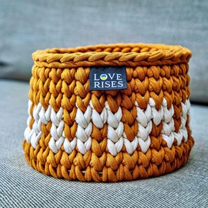 Peut inclure: Panier rond tricoté à la main, jaune moutarde et blanc. Il présente un motif en chevrons répété et une étiquette « LOVE RISES ». Fabriqué en fil épais et texturé, il est idéal pour le rangement.