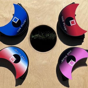 May include: Four half-moon shaped game pieces with a gradient design. The pieces are blue, red, pink, and red and white. Each piece has a black circle with a white square in the center and the text "THE PLUG" written around the circle.
