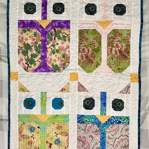 May include: A rectangular quilt featuring four stylized owl figures arranged in a grid. Each owl has white quilted fabric, dark peacock-feather patterned eyes, and bodies made from various floral and paisley print fabrics in purple, green, and blue.