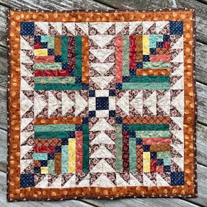 Puede incluir: Una colcha de tela cuadrada que presenta un cuadrado central azul oscuro rodeado por un patrón geométrico de triángulos y tiras rectangulares en tonos rojo, verde, amarillo y crema con estampados florales y de cachemira, con un borde de bellotas marrones.